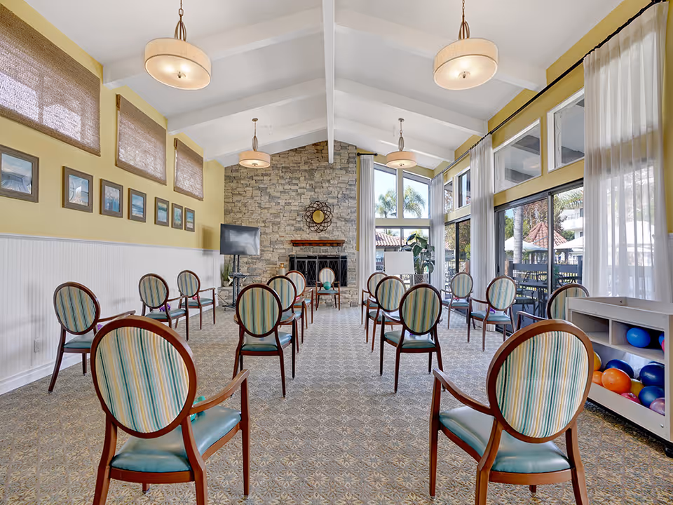 A spacious room with high vaulted ceilings and large windows letting in natural light. The room is arranged with multiple wooden chairs with striped upholstery spaced apart, facing a stone fireplace and a TV on a stand. The walls are painted yellow with white wainscoting, and there are framed pictures and woven window shades on one side. On the right, there is a shelf with colorful balls. Outside the windows, palm trees and a patio area are visible.