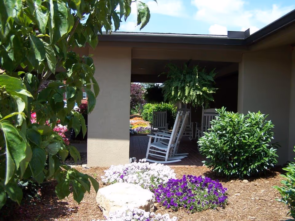 Outdoor patio area with white rocking chairs under a covered porch surrounded by green bushes, hanging fern plants, and colorful flower beds with purple and white flowers.