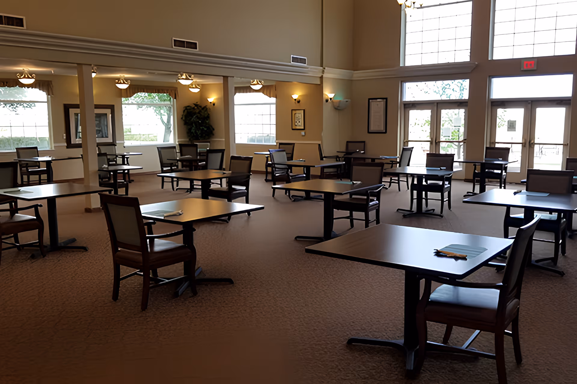 A spacious dining room with multiple square tables and chairs arranged neatly. The room has large windows allowing natural light to fill the space, beige walls, and carpeted flooring. Each table has a placemat and a napkin, and there are wall-mounted lights and framed pictures on the walls.