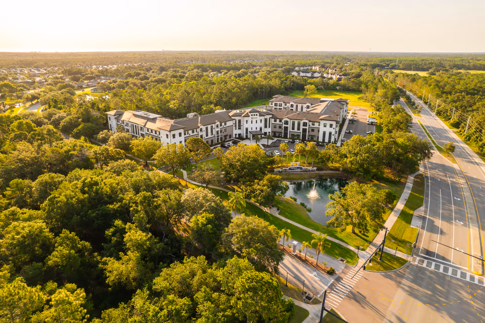 Aerial view of a senior living facility named Experience Senior Living surrounded by lush green trees and landscaping, with a pond featuring a fountain in front of the building. The facility is adjacent to a multi-lane road with sidewalks and crosswalks.