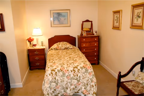 Small bedroom with a single bed covered in a floral bedspread, a bedside table with a lamp, a wooden dresser, and framed pictures on the walls.