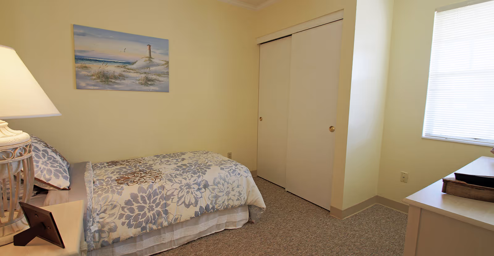 A small bedroom with a single bed covered in a floral patterned comforter. There is a nightstand with a lamp and a picture frame next to the bed. A painting of a lighthouse on a beach hangs on the wall above the bed. The room has a window with blinds and a closet with sliding doors.