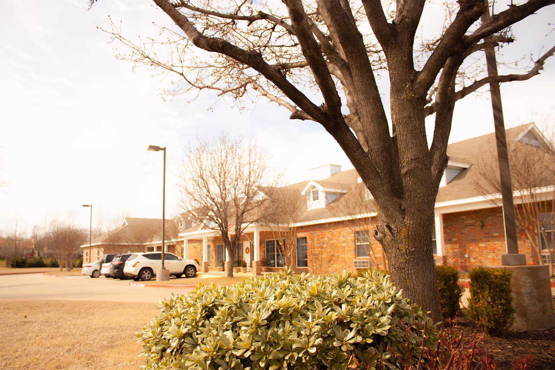 Exterior view of a single-story brick building with a sloped roof, surrounded by leafless trees and bushes. Several cars are parked in the parking lot adjacent to the building under a partly cloudy sky.