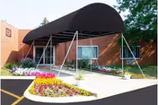 Entrance of LakePointe Senior Care & Rehab Center with a black canopy over the walkway, flower beds with colorful flowers on either side, and a brick building in the background.