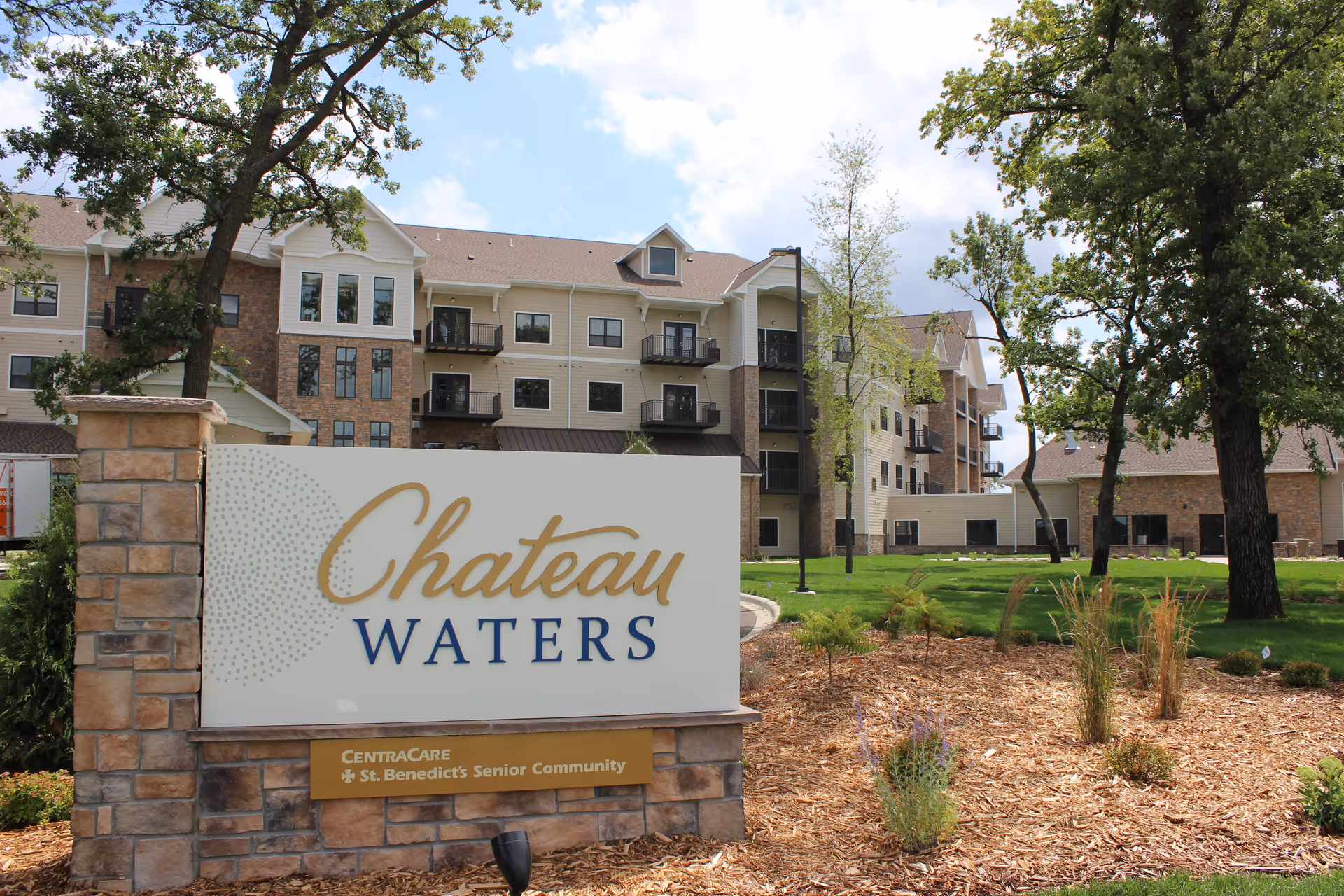 Exterior view of Chateau Waters, a senior living community building with multiple floors, balconies, and a landscaped garden area with trees and plants. A large sign in front reads 'Chateau Waters' with additional text 'CentraCare St. Benedict's Senior Community'.