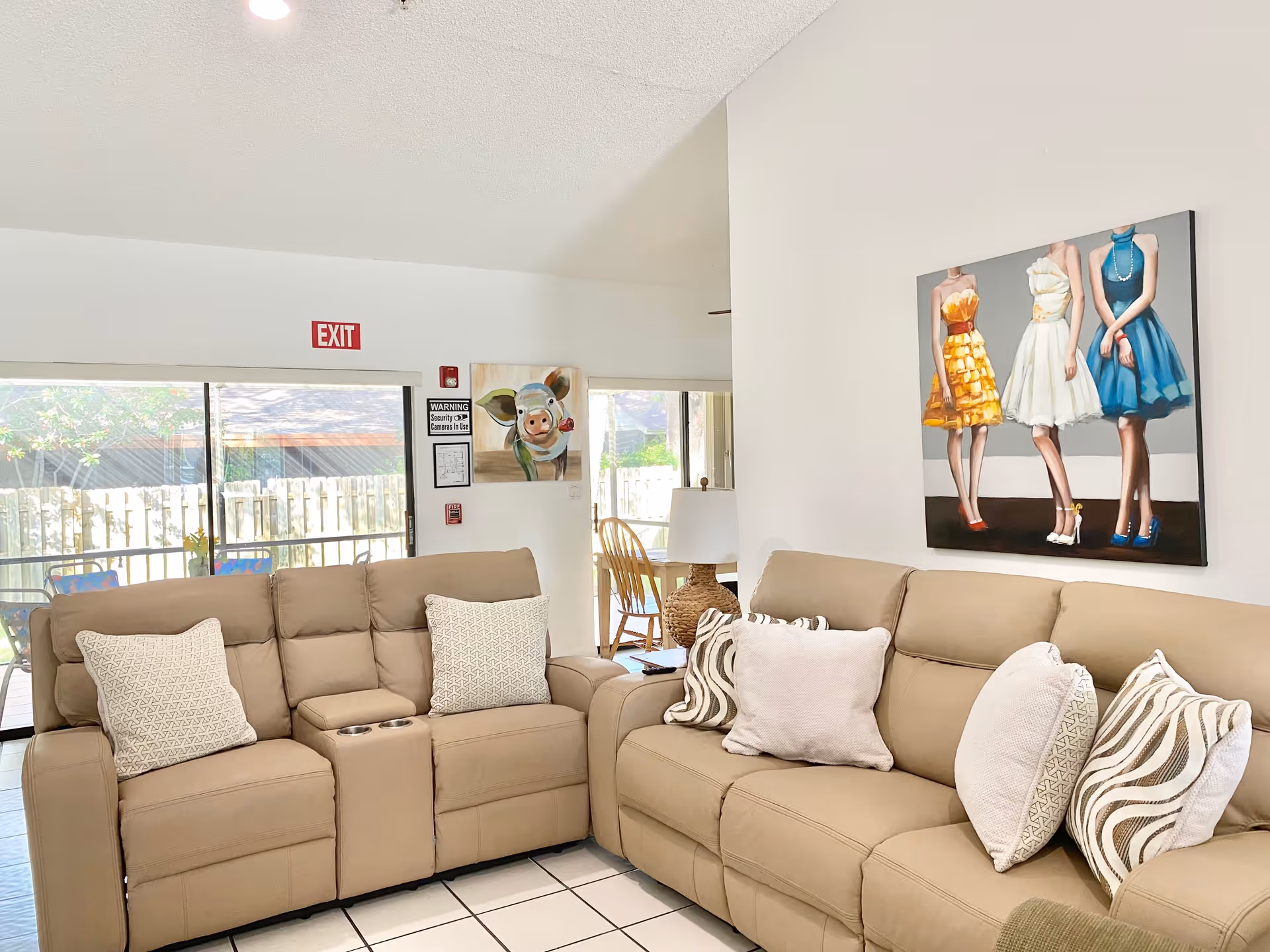 A bright living room with two beige sofas adorned with patterned throw pillows. On the wall above one sofa is a painting of three women in colorful dresses. A sliding glass door with an exit sign above it leads to an outdoor area with a wooden fence and patio furniture. A painting of a pig and a warning sign about security cameras are visible on the wall near the door. A wicker lamp and wooden chair are also present in the background.