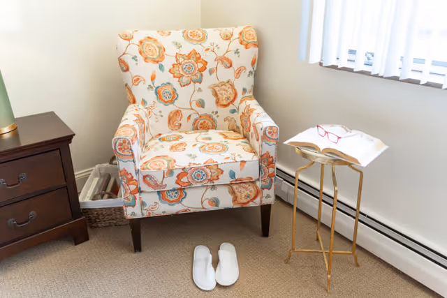 A cozy corner of a room featuring a floral patterned armchair with orange, blue, and beige tones. In front of the chair is a pair of white slippers on a beige carpet. To the left of the chair is a dark wooden nightstand with two drawers and a green lamp on top. Behind the chair is a woven basket with rolled magazines or papers. To the right of the chair is a small gold-colored side table shaped like an open book holding an open book and a pair of red reading glasses. A window with vertical blinds lets in natural light from the right side.