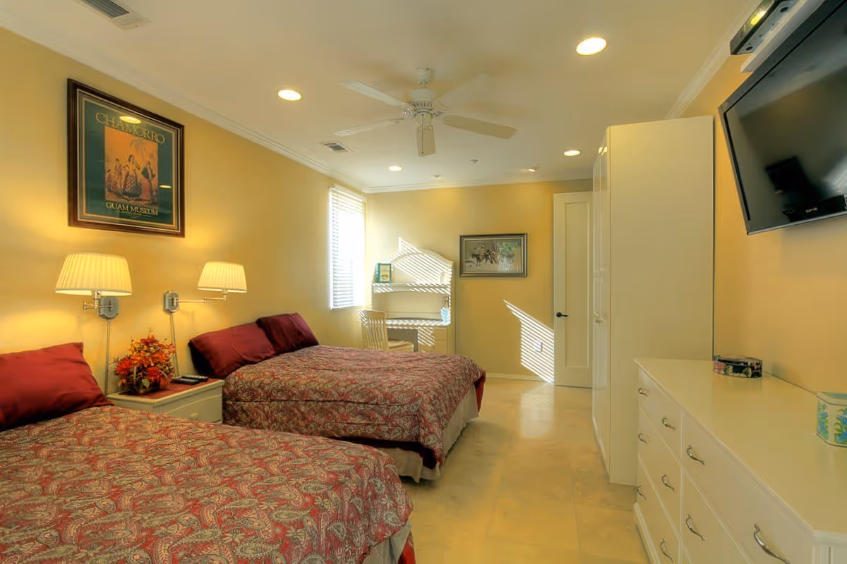 A well-lit bedroom with two beds covered in red patterned bedspreads, each with a matching pillow. Between the beds is a nightstand with a floral arrangement and two wall-mounted lamps above it. On the right side of the room is a white dresser with multiple drawers and a flat-screen TV mounted above it. At the far end of the room, there is a small desk with a chair positioned near a window with blinds. The walls are painted a soft yellow, and there are two framed pictures hanging on the walls. The floor is tiled, and a ceiling fan with lights is mounted on the ceiling.