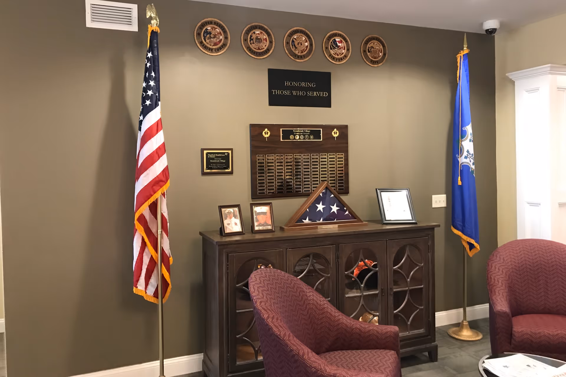 A memorial display in a room with two maroon armchairs. The display includes an American flag and a Connecticut state flag on stands, a wooden cabinet with a folded American flag in a triangular case, framed photographs, a plaque, and military service emblems mounted on the wall above a sign that reads 'Honoring Those Who Served.'