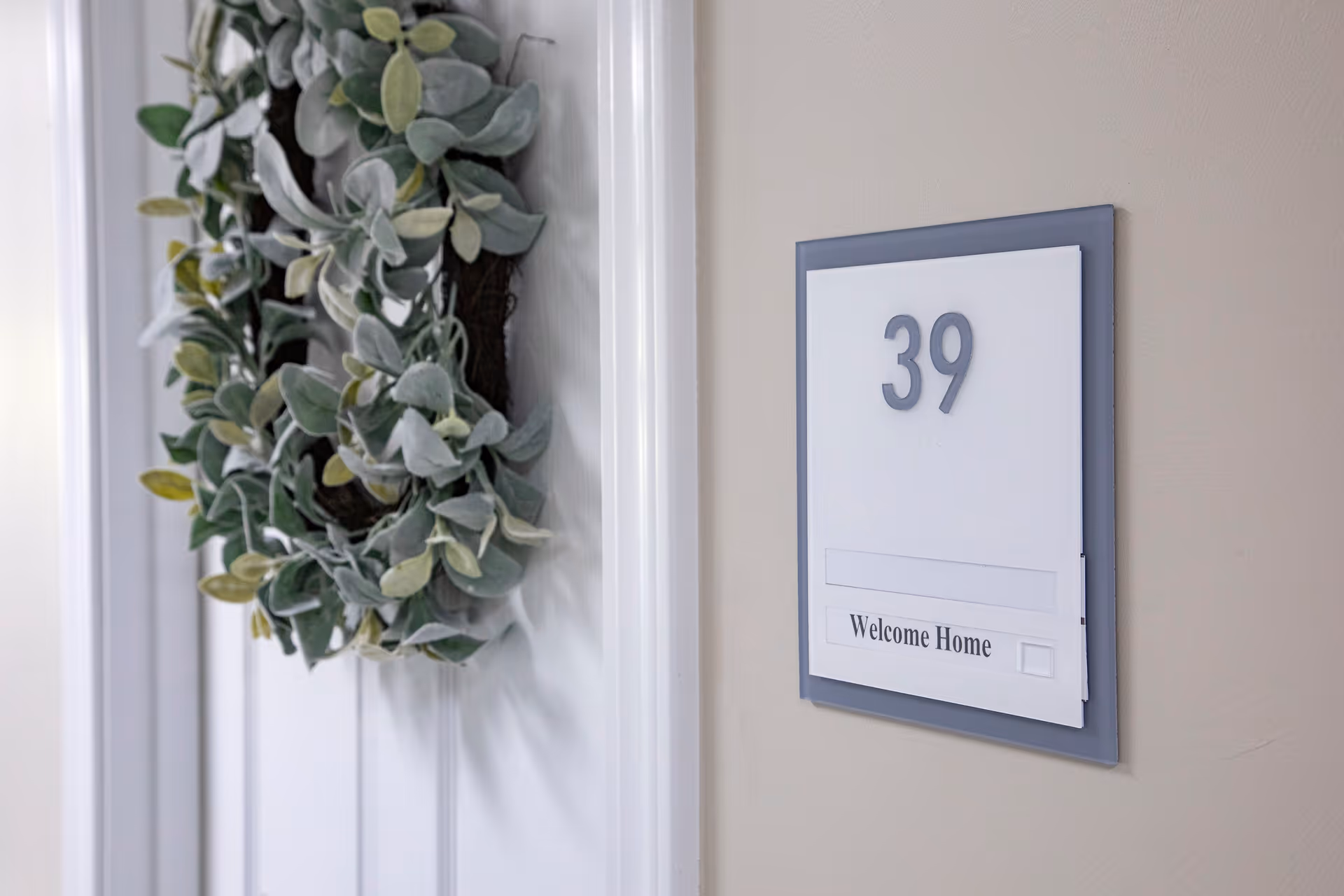 Close-up view of a white door with a decorative green leafy wreath hanging on it. Next to the door, a wall-mounted sign displays the number 39 and the words 'Welcome Home'.