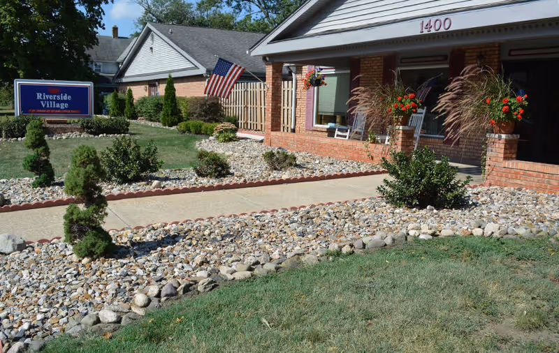 Exterior view of Riverside Village senior living facility showing a brick building with a covered porch, two white rocking chairs, hanging flower baskets, an American flag, and a landscaped front yard with rocks, small bushes, and a concrete walkway.