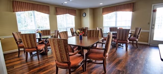 Dining room with several wooden tables and upholstered chairs, hardwood floors, large windows with valances, and small floral centerpieces.