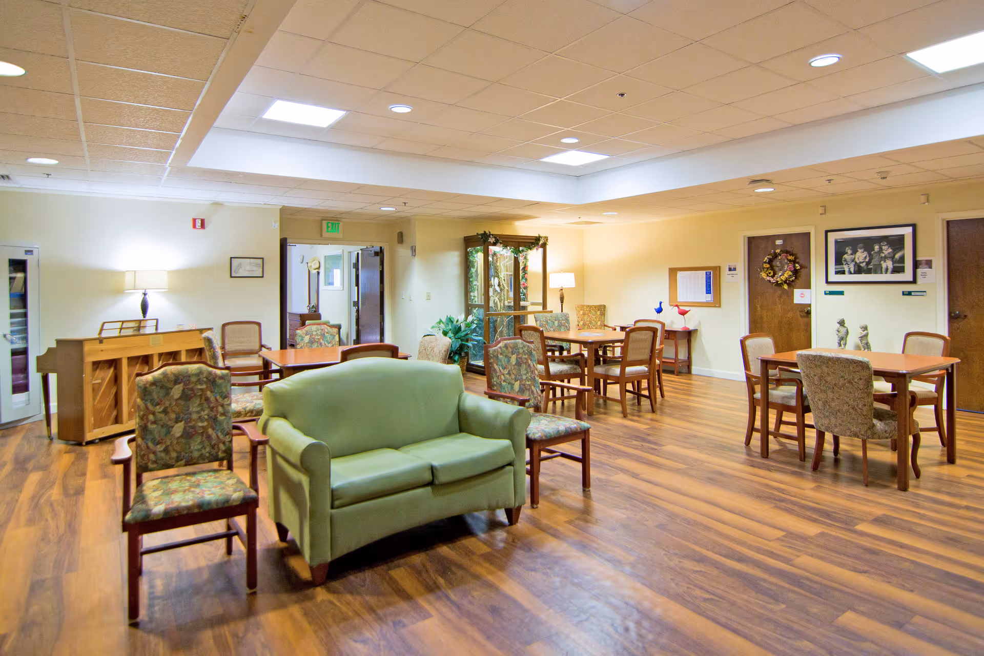 A spacious common area in a senior living facility with wooden floors, several tables and chairs arranged for group seating, a green loveseat, floral upholstered chairs, a piano against the wall, and soft lighting from ceiling fixtures and lamps. The walls are decorated with framed pictures and a wreath on a door.