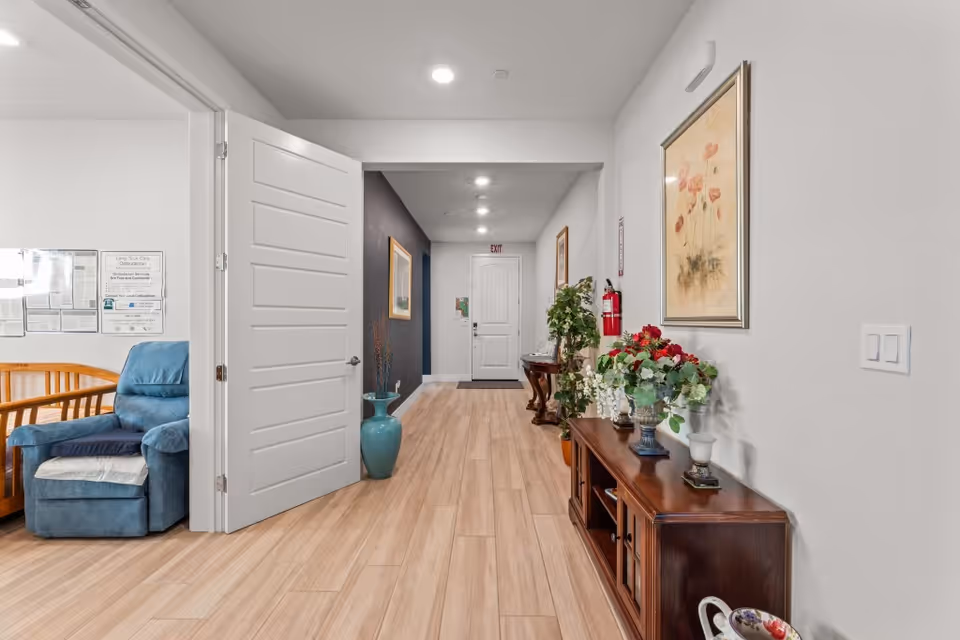 A well-lit hallway in a senior living facility with light wood flooring and white walls. On the right side, there is a wooden console table decorated with flower arrangements and a framed floral painting above it. A fire extinguisher is mounted on the wall. On the left side, an open door reveals a room with a blue recliner chair and a wooden bench. The hallway leads to a white exit door at the end, with framed pictures and a large blue vase along the walls.