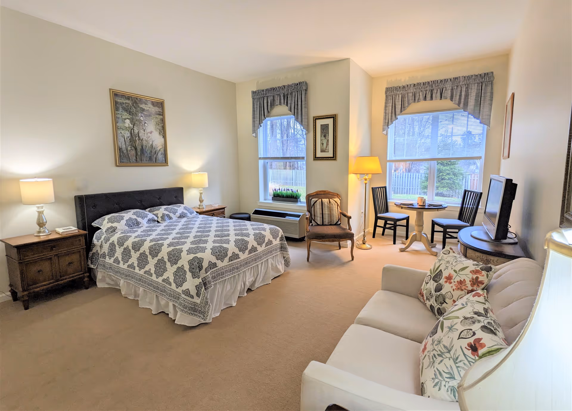 A bright and cozy bedroom in a senior living facility featuring a large bed with patterned bedding, two bedside tables with lamps, a cushioned armchair, a small round table with two chairs near two windows with valances, a TV on a stand, and a white sofa with floral pillows.