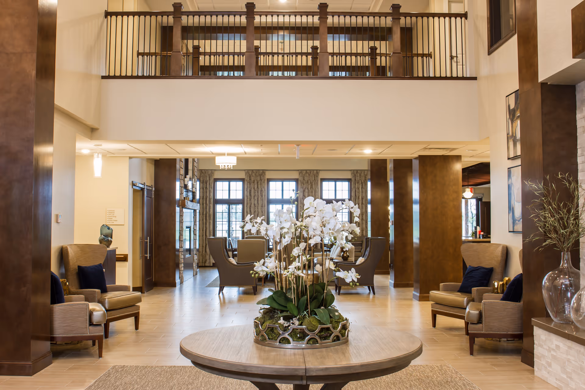 Spacious and elegant senior living facility common area with a round wooden table in the foreground holding a large floral arrangement of white orchids. The room features high ceilings with a balcony railing above, multiple comfortable armchairs with dark cushions, large windows with patterned curtains, and warm lighting. The decor includes wooden pillars and modern artwork on the walls.