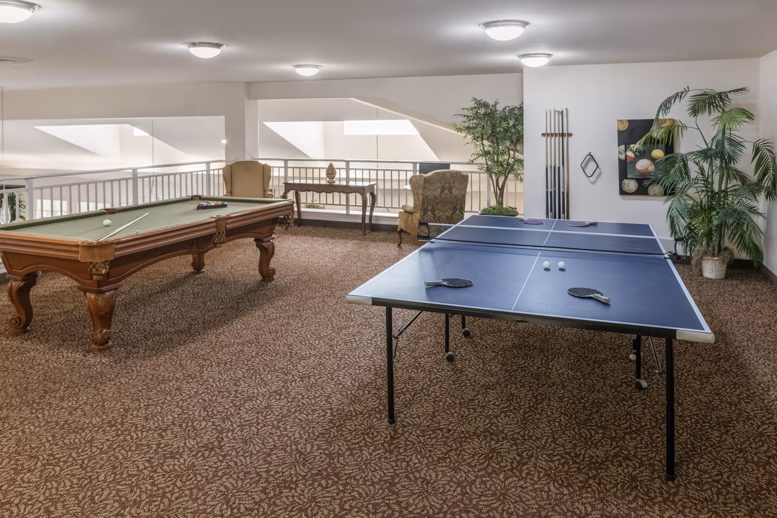 A recreational room with a pool table and a ping pong table. The room has patterned carpet, two armchairs, a small table, and several potted plants. Pool cues are mounted on the wall along with a decorative picture of billiard balls.