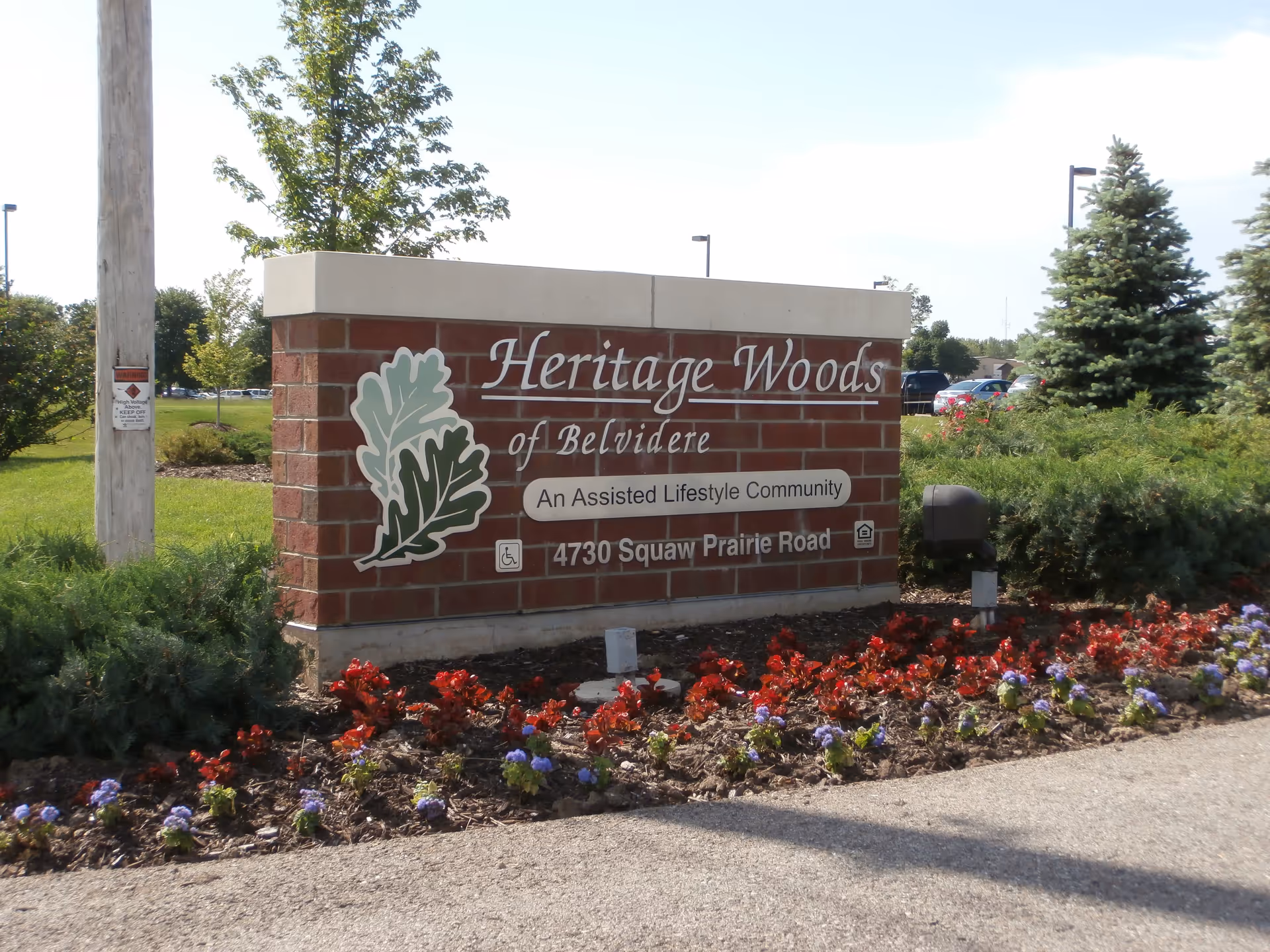 A brick sign for Heritage Woods of Belvidere, an assisted lifestyle community, located at 4730 Squaw Prairie Road. The sign is surrounded by flower beds with red and purple flowers, green shrubs, and trees in the background under a clear sky.