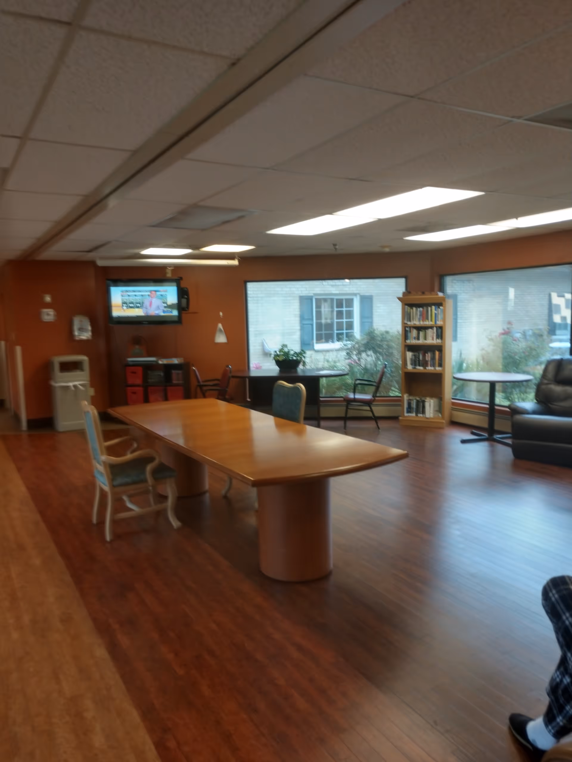 A common area in a senior living facility with a large wooden table and chairs, a bookshelf filled with books, a television mounted on the wall, large windows showing an outside view, and a comfortable black recliner chair.