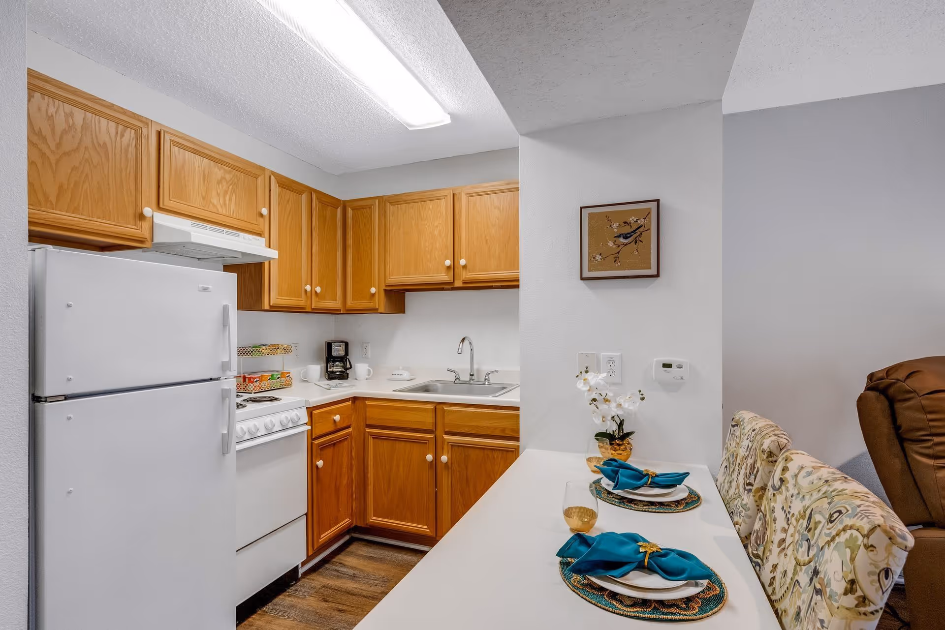 Small kitchenette with a white refrigerator and stove, wooden cabinets, sink, and a countertop set with two place settings.