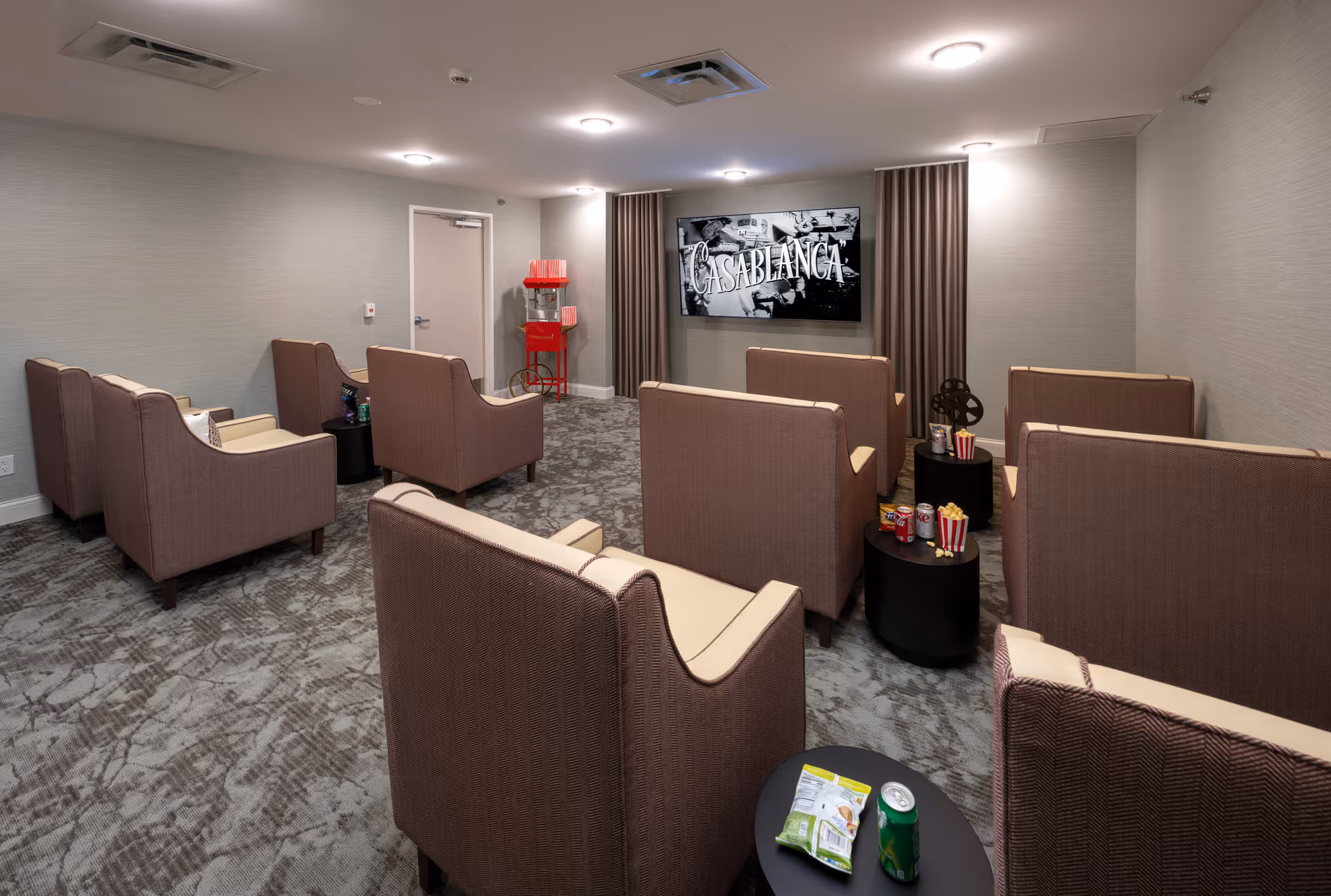 A small movie theater room with several rows of brown cushioned chairs facing a large screen displaying the movie Casablanca. There are small black tables between the chairs with snacks and drinks, and a red popcorn machine is visible in the back corner of the room.