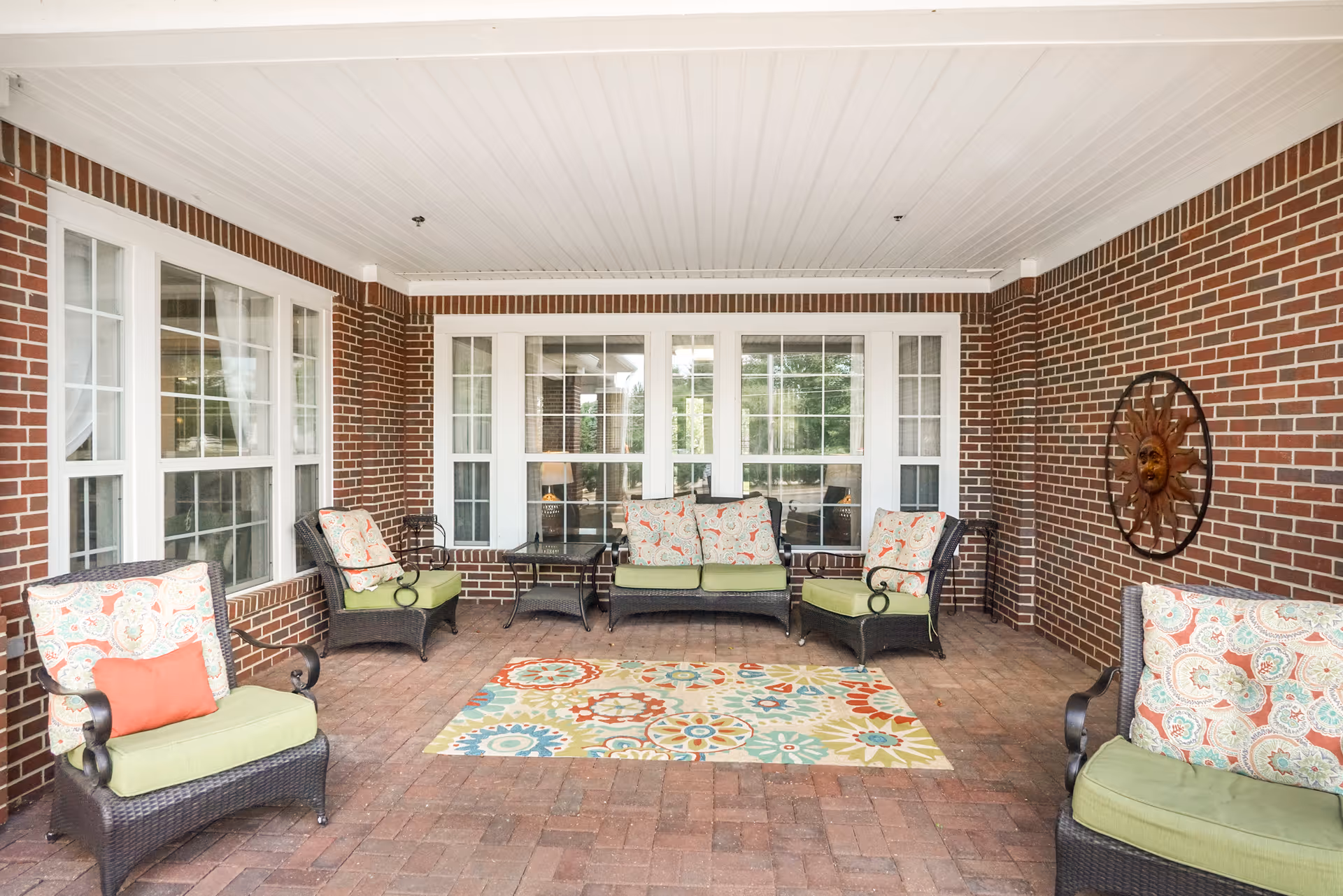 Covered outdoor patio area with brick walls and floor, featuring wicker furniture with green cushions and patterned pillows, a colorful patterned rug, and a decorative sun face wall hanging.