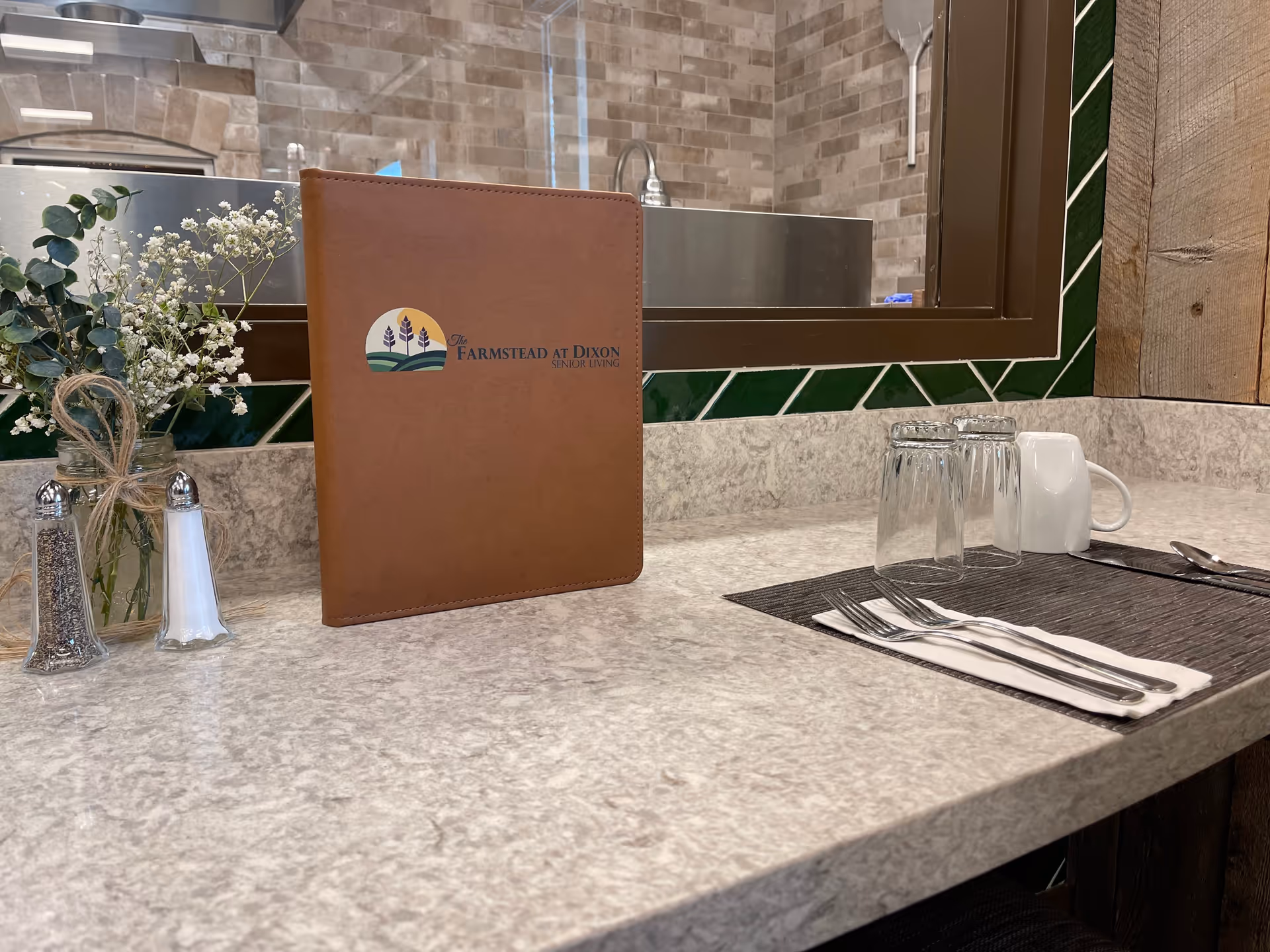 Countertop dining area with a brown "The Farmstead at Dixon" menu folder, glasses, utensils, salt and pepper shakers, and a small floral arrangement in front of a pass-through window to the kitchen.