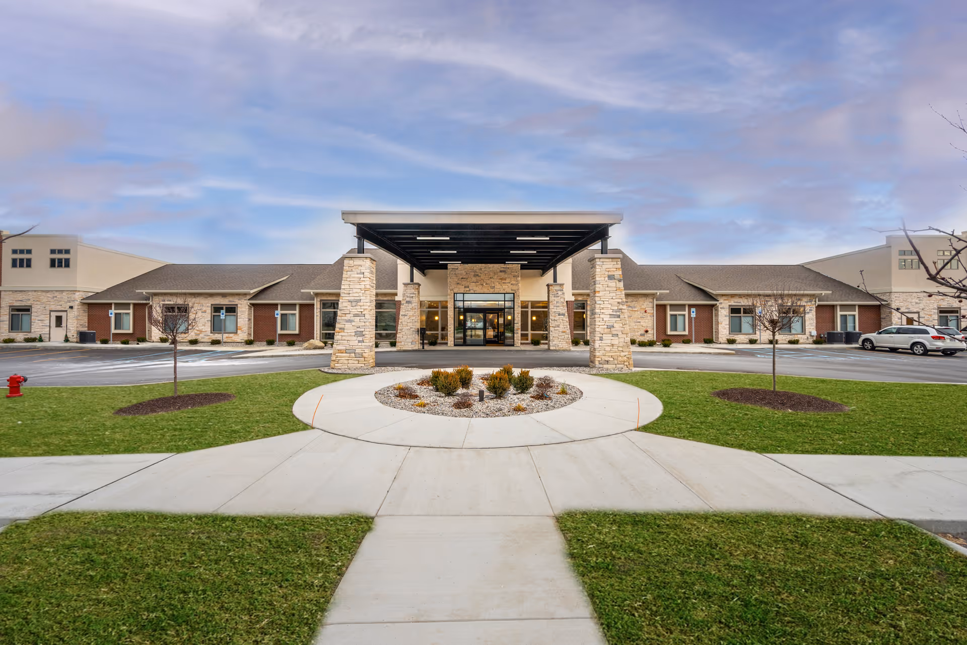 Front exterior view of Hampton Manor of Commerce building with a covered entrance supported by stone pillars, surrounded by a circular driveway, green lawn, and a partly cloudy sky.