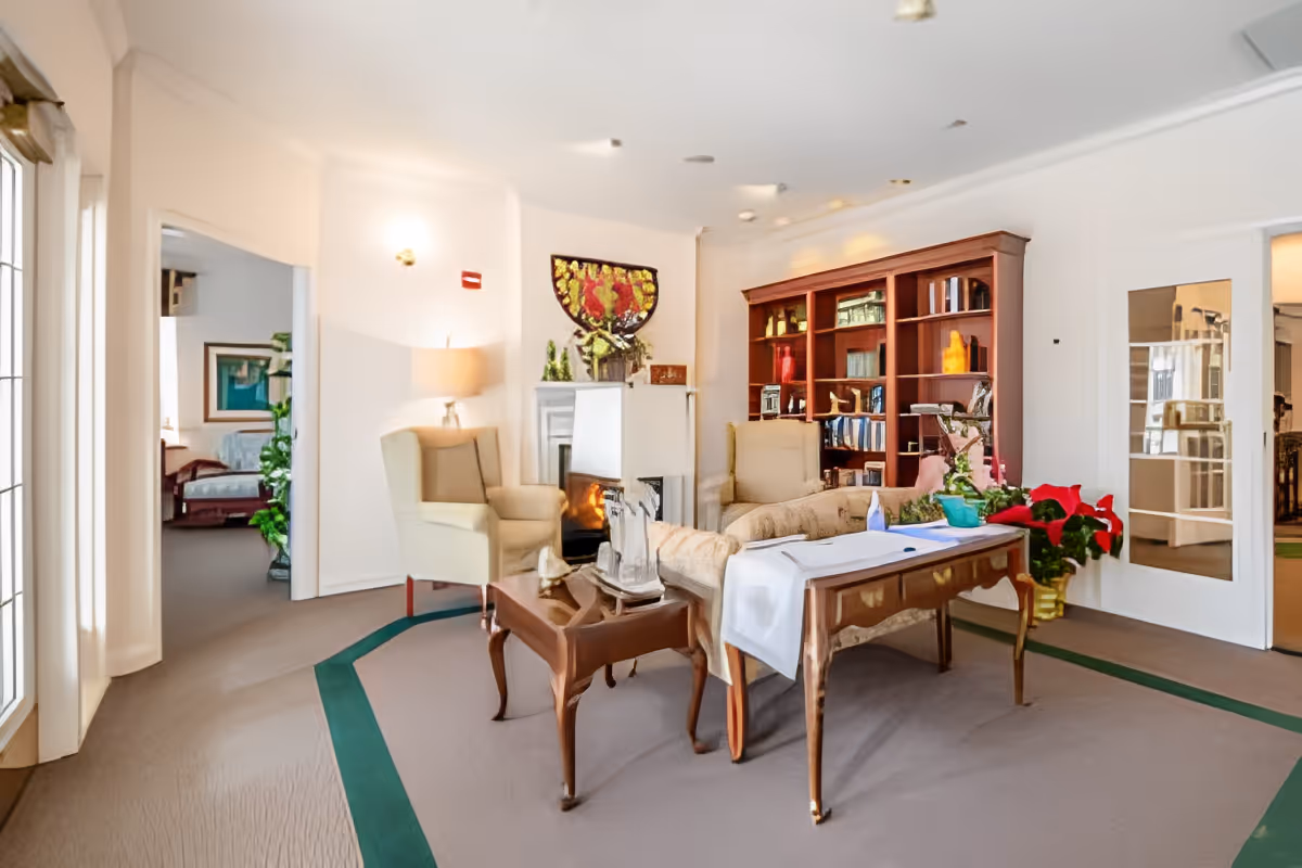 Bright sitting room with armchairs, a sofa, a wooden bookshelf, fireplace, and a table with a tablecloth.
