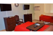 Interior view of a nursing facility room with two beds covered in red blankets, a wooden chair, two dressers, a wall-mounted TV, an air conditioning unit, and a window with blinds.
