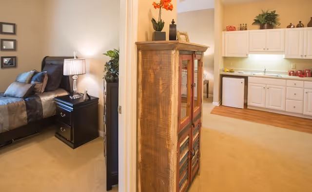 Interior view of a senior living facility showing a bedroom with a bed, pillows, a nightstand with a lamp, and a small kitchen area with white cabinets, a sink, and a mini refrigerator. A wooden cabinet with decorative items on top is positioned between the bedroom and kitchen area.