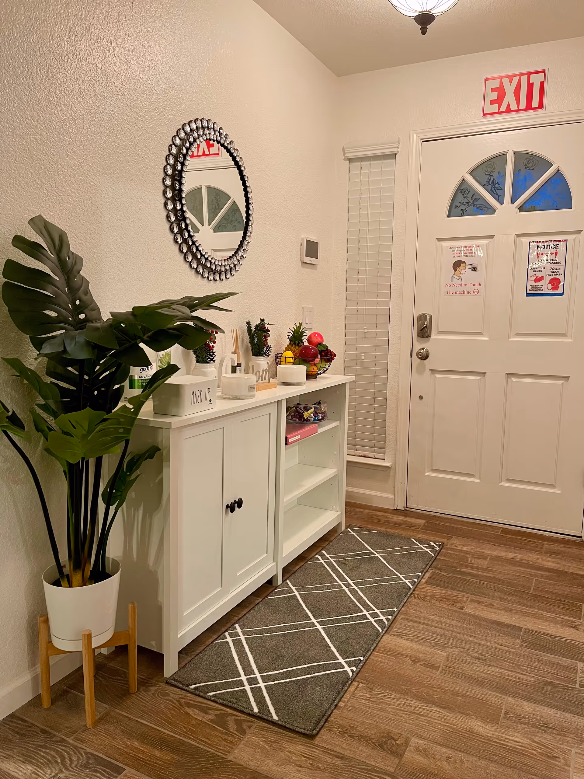 Entryway area with a white front door featuring a semi-circular window with floral designs and an EXIT sign above. To the left of the door is a white cabinet with various decorative items including a fruit basket, small plants, and containers. A round mirror with a decorative frame hangs above the cabinet. A tall green potted plant stands on a wooden stand next to the cabinet. The floor is wood with a gray and white patterned runner rug in front of the cabinet and door.