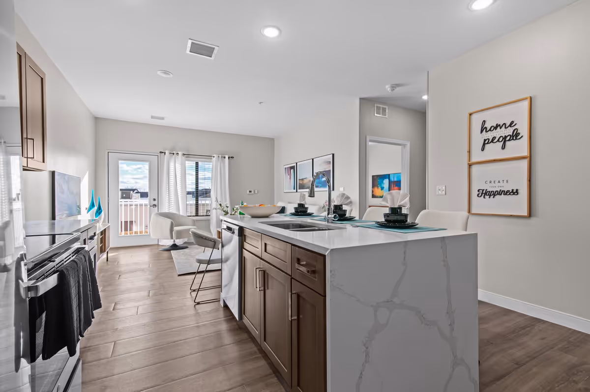 Modern kitchen and living area with a large marble kitchen island featuring a sink and place settings. The room has wooden floors, light gray walls, and a door with a window leading to an outdoor balcony. There are two framed inspirational quotes on the wall and contemporary artwork visible in an adjacent room.