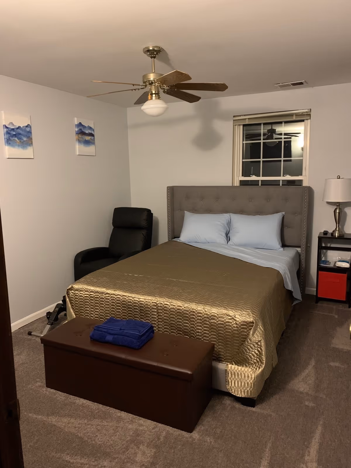 A bedroom with a bed covered in a gold quilt and light blue pillows. There is a brown storage bench at the foot of the bed with folded blue towels on top. A black recliner chair is placed in the corner near two blue and white abstract paintings on the wall. A ceiling fan with wooden blades and a light fixture hangs from the ceiling. A window with white blinds is behind the bed, and a small black nightstand with a lamp and some items is next to the bed.