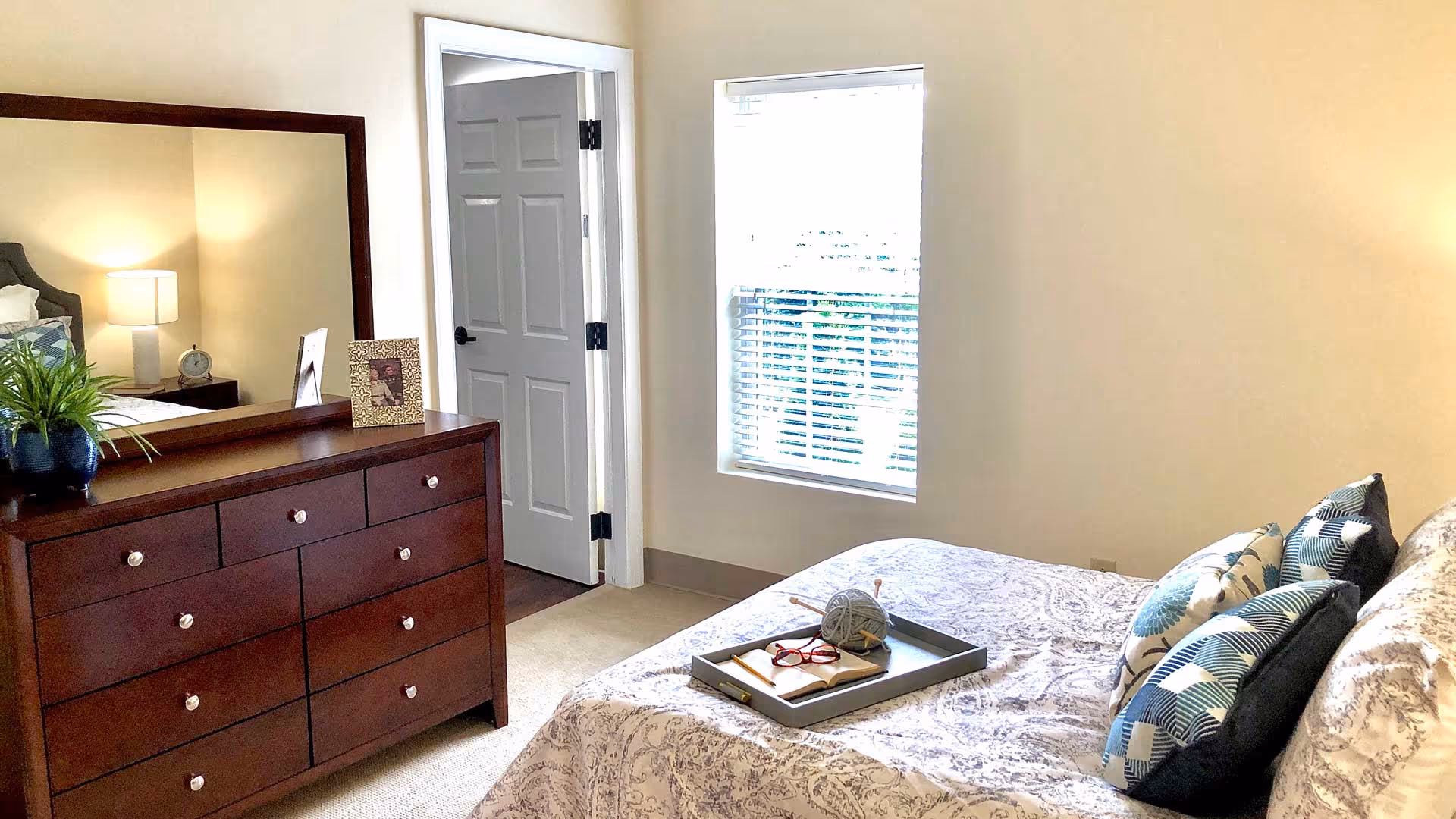 A bright bedroom with a bed covered in patterned bedding and several decorative pillows. A tray on the bed holds a ball of yarn with knitting needles, a pair of red glasses, and an open book. To the left, there is a dark wooden dresser with a large mirror, a potted plant, and framed photos. A door is open next to a window with white blinds, letting in natural light.