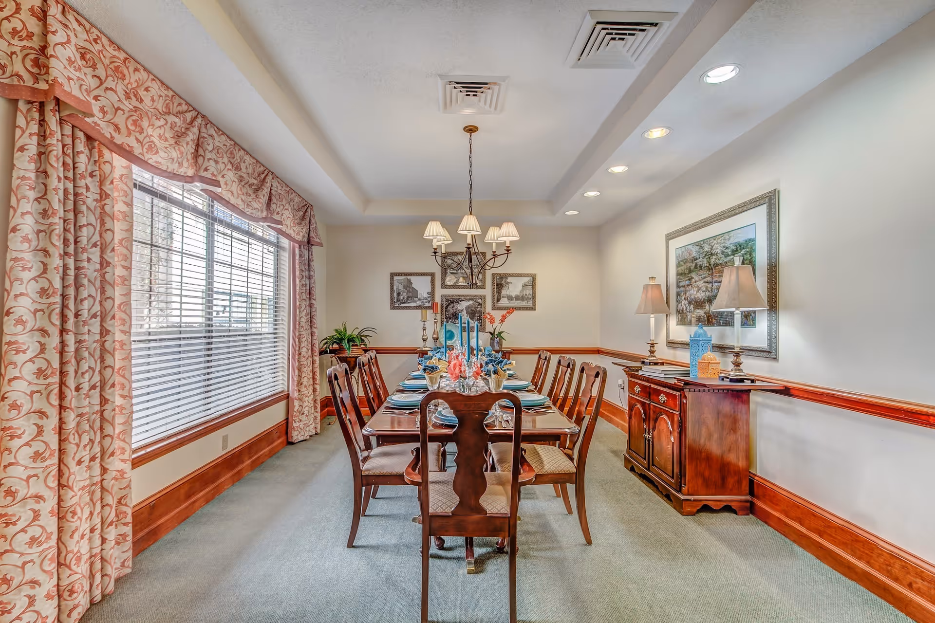A formal dining room with a long wooden dining table set for eight people. The table is decorated with blue candles and floral arrangements. There are eight wooden chairs around the table. The room has large windows with patterned curtains on the left side, a chandelier hanging from the ceiling, framed pictures on the far wall, and a wooden sideboard with two lamps and decorative items on the right side.