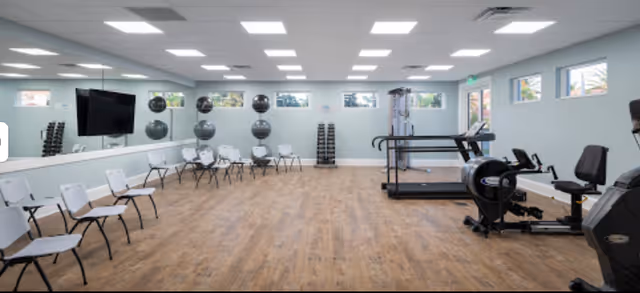 A spacious fitness room with wooden flooring, white walls, and multiple ceiling lights. The room features a row of white chairs along one wall, a large mirror, exercise balls stacked on racks, dumbbells, a treadmill, and other exercise equipment. Small windows near the ceiling allow natural light to enter.