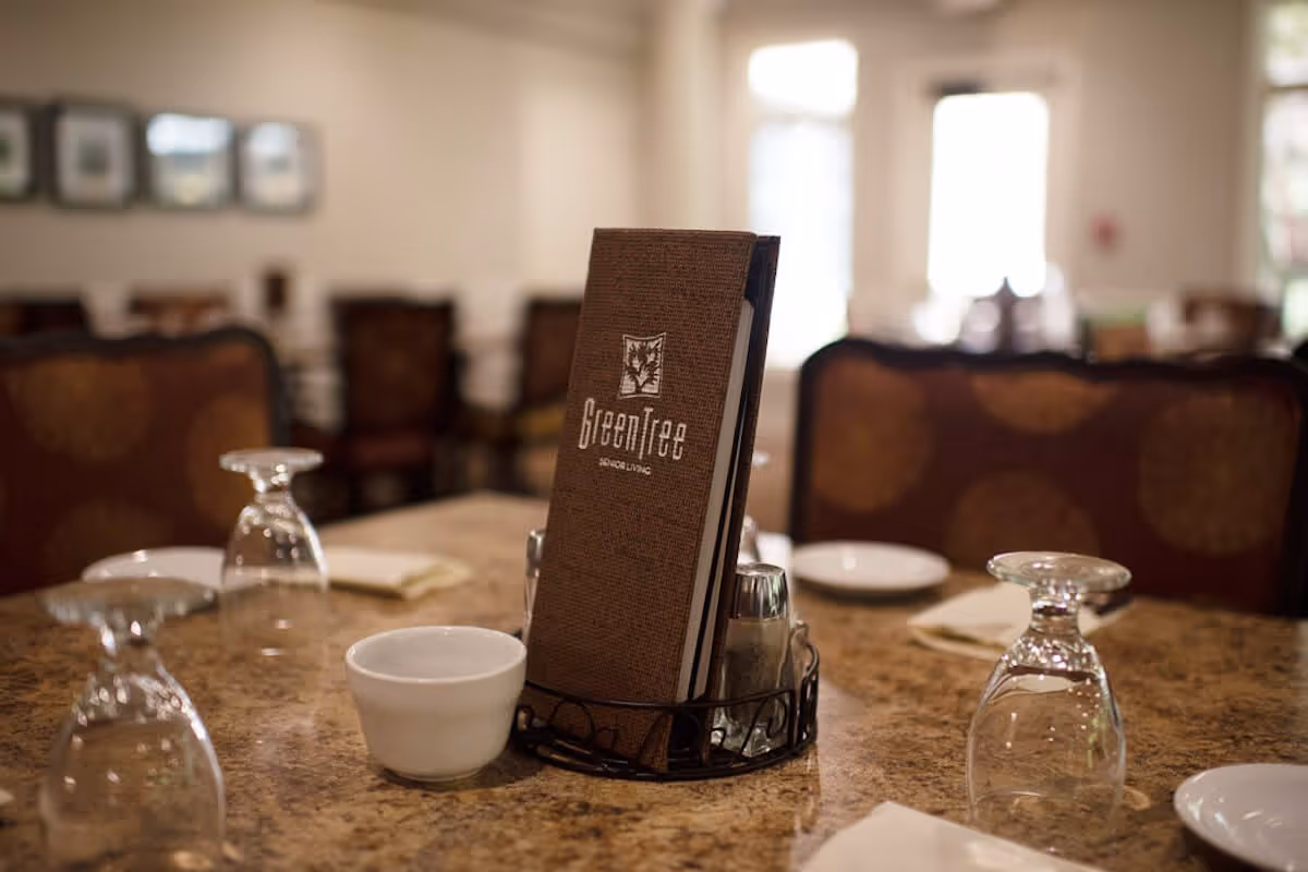 A dining table set with upside-down wine glasses, white bowls, plates, and napkins. In the center of the table is a menu holder with a menu labeled 'GreenTree Senior Living'. The background shows upholstered chairs and a softly lit dining room.