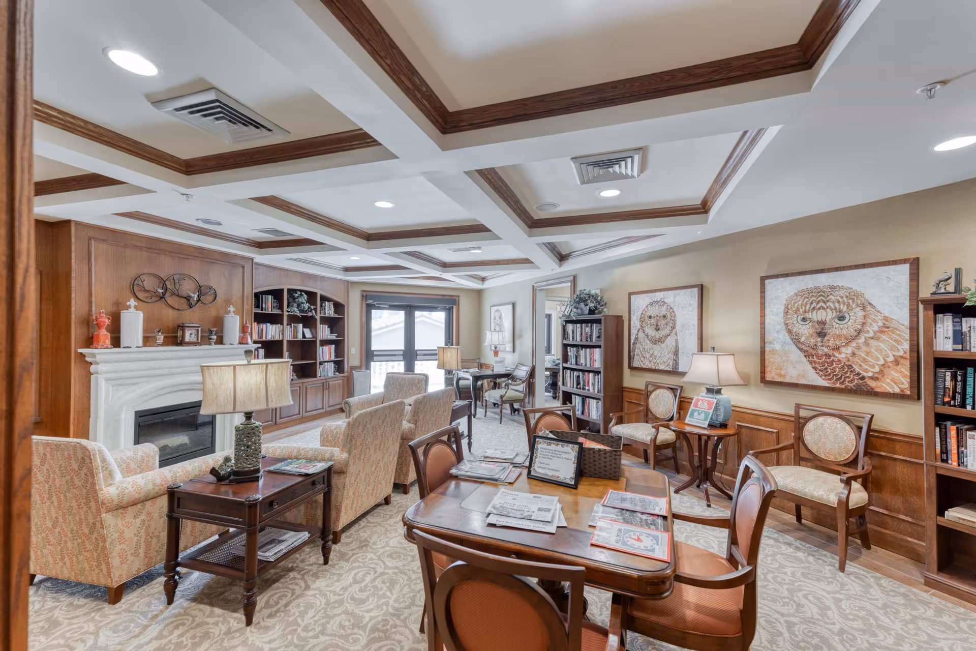 A cozy senior living facility common area with patterned armchairs, wooden tables, and bookshelves. The room features a white fireplace with decorative items on the mantel, two large owl paintings on the wall, and a ceiling with wooden beams and recessed lighting. There are newspapers and informational materials on the tables, and a window with a view outside.