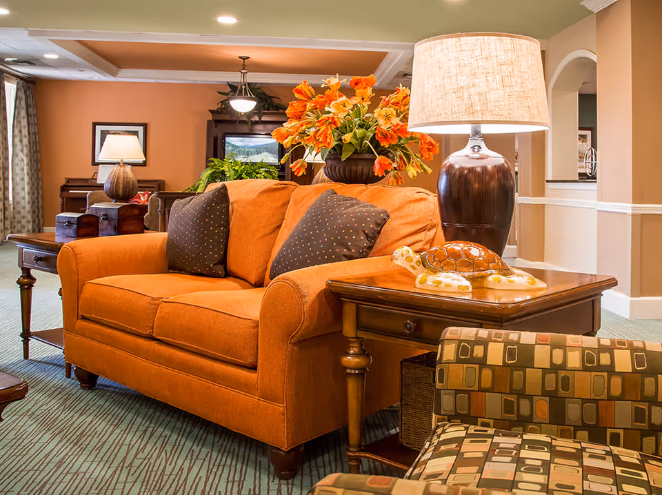A cozy living room area featuring an orange loveseat with two dark patterned pillows, a wooden side table with a lamp and a decorative turtle figurine, a patterned armchair in the foreground, and a floral arrangement in the background. The room has warm beige walls and soft lighting.