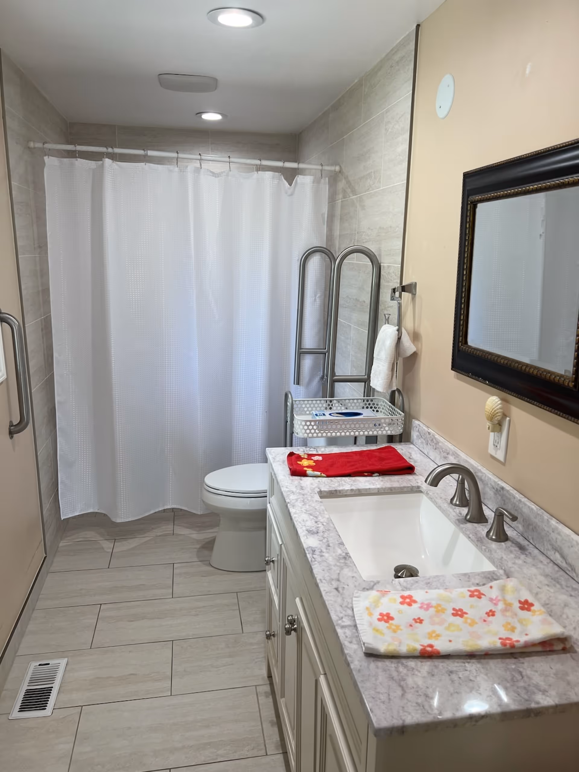 A clean and modern bathroom with a white shower curtain, a toilet, a white vanity with a marble countertop, a sink with silver faucets, a mirror with a dark frame, and two folded towels on the countertop. The walls are tiled and there are two ceiling lights.