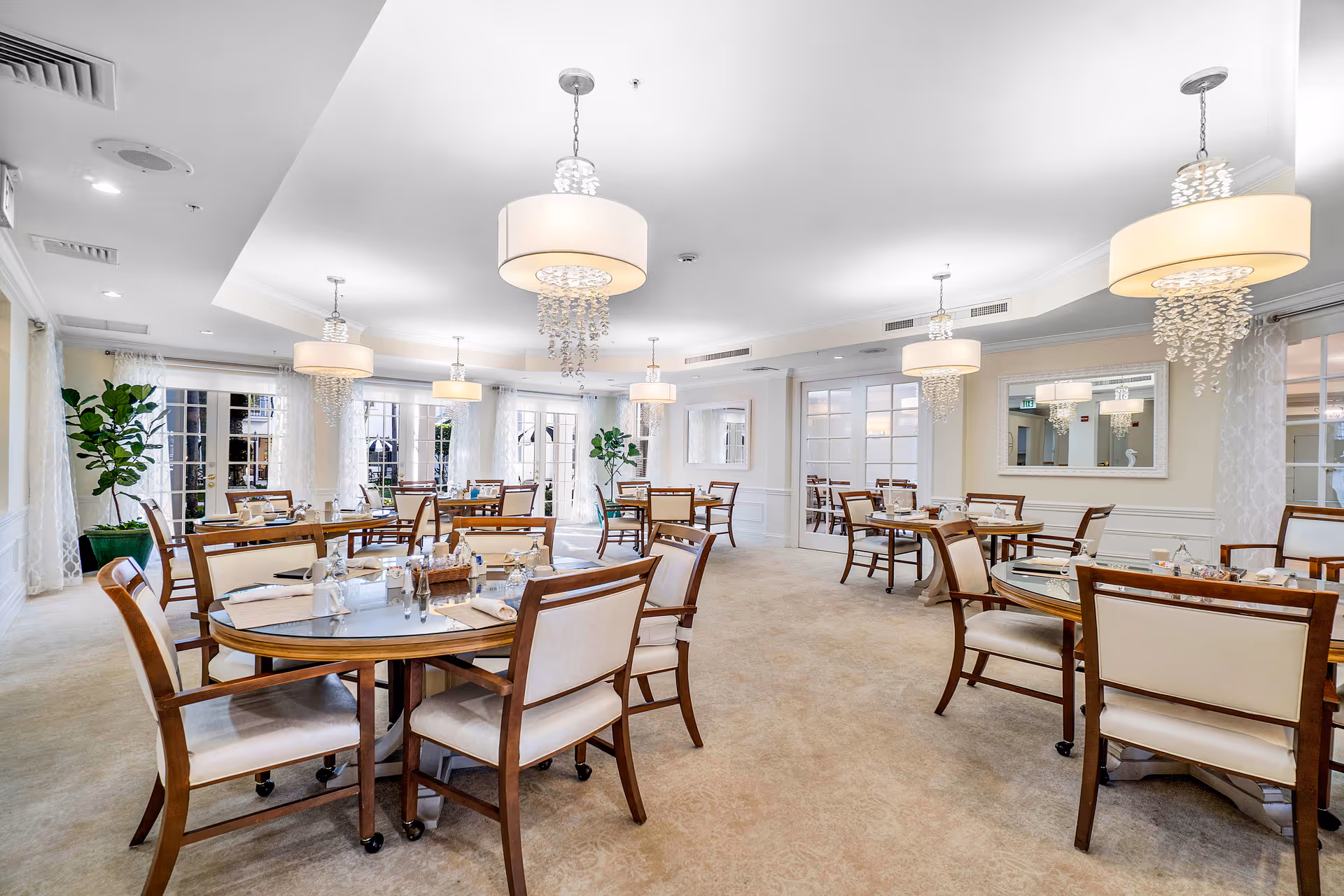 Bright and elegant dining room with multiple round tables set with glassware, napkins, and condiments. The room features beige carpeting, cream-colored walls, large windows with sheer curtains, decorative mirrors, and modern chandeliers with hanging crystal accents. Several wooden chairs with cream cushions surround each table.