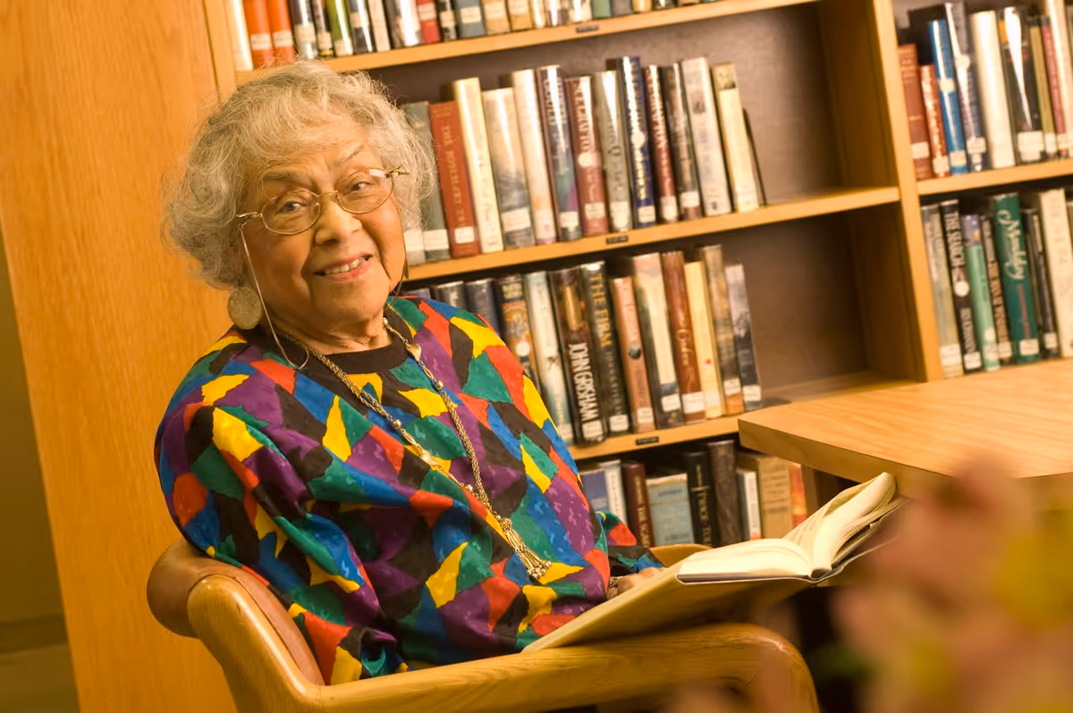 An elderly woman with gray hair and glasses is sitting in a wooden chair in a library or reading room. She is wearing a colorful patterned sweater and smiling while holding an open book. Behind her are shelves filled with books.
