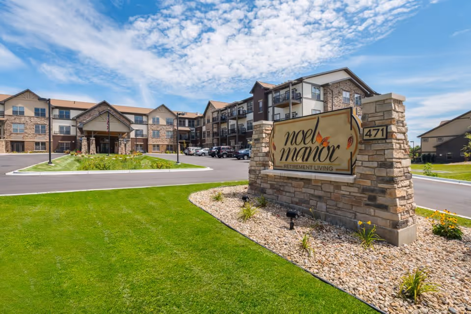 Exterior view of Noel Manor Retirement Living facility showing a large three-story building with stone and siding facade, a driveway, parked cars, and a landscaped lawn with a stone sign displaying the facility name and address number 471 under a partly cloudy blue sky.