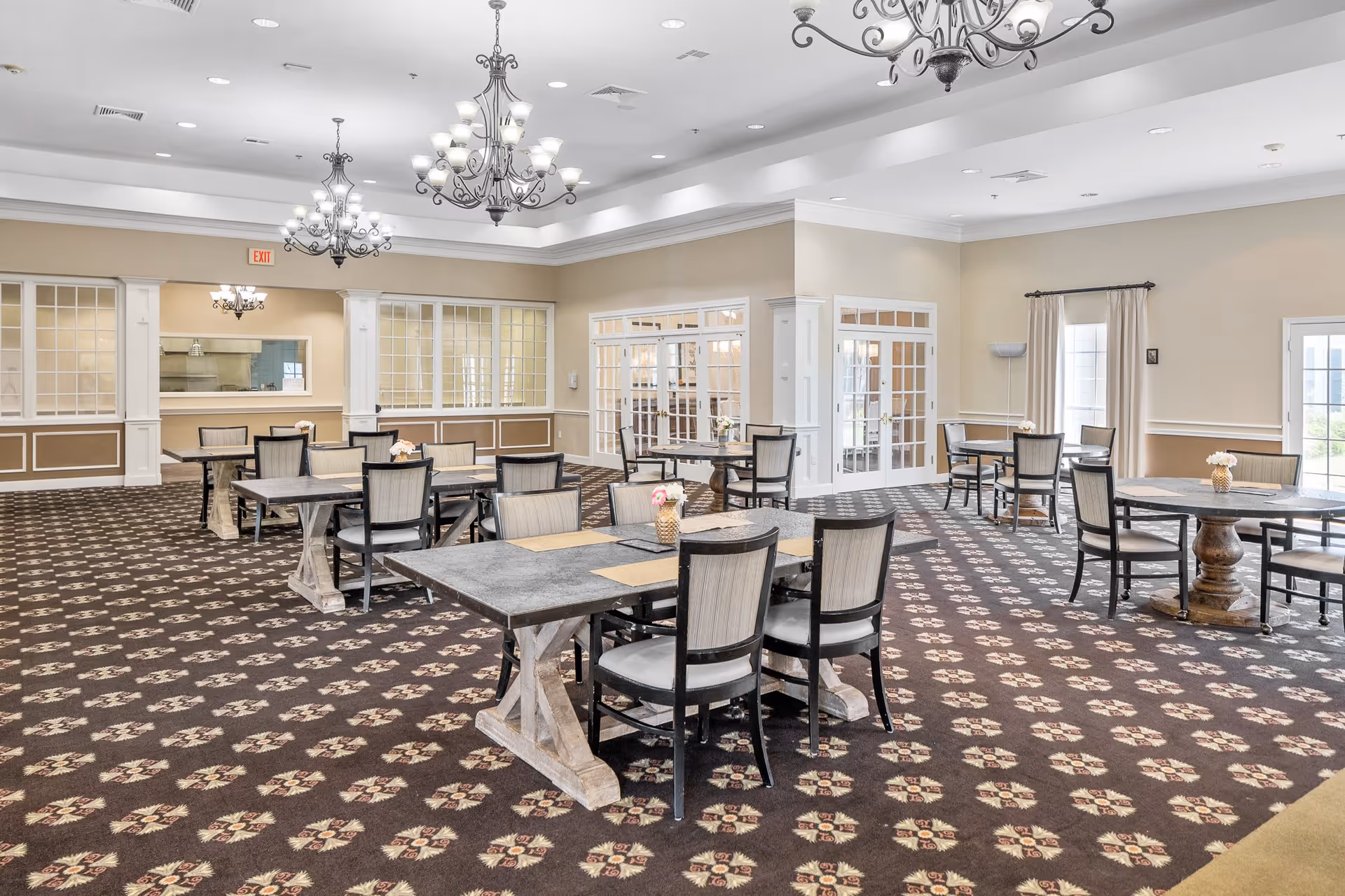 A spacious dining room with multiple tables and chairs arranged neatly. The room features patterned carpet flooring, beige walls with white trim, large windows with curtains, and elegant chandeliers hanging from the ceiling. There is a serving window on one side and glass double doors leading to another area.