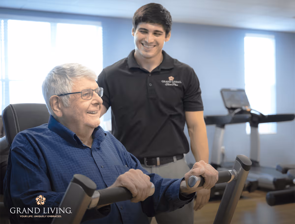 An elderly man using an exercise machine in a fitness room while a younger man, wearing a black polo shirt with the Grand Living logo, stands beside him smiling and offering support.