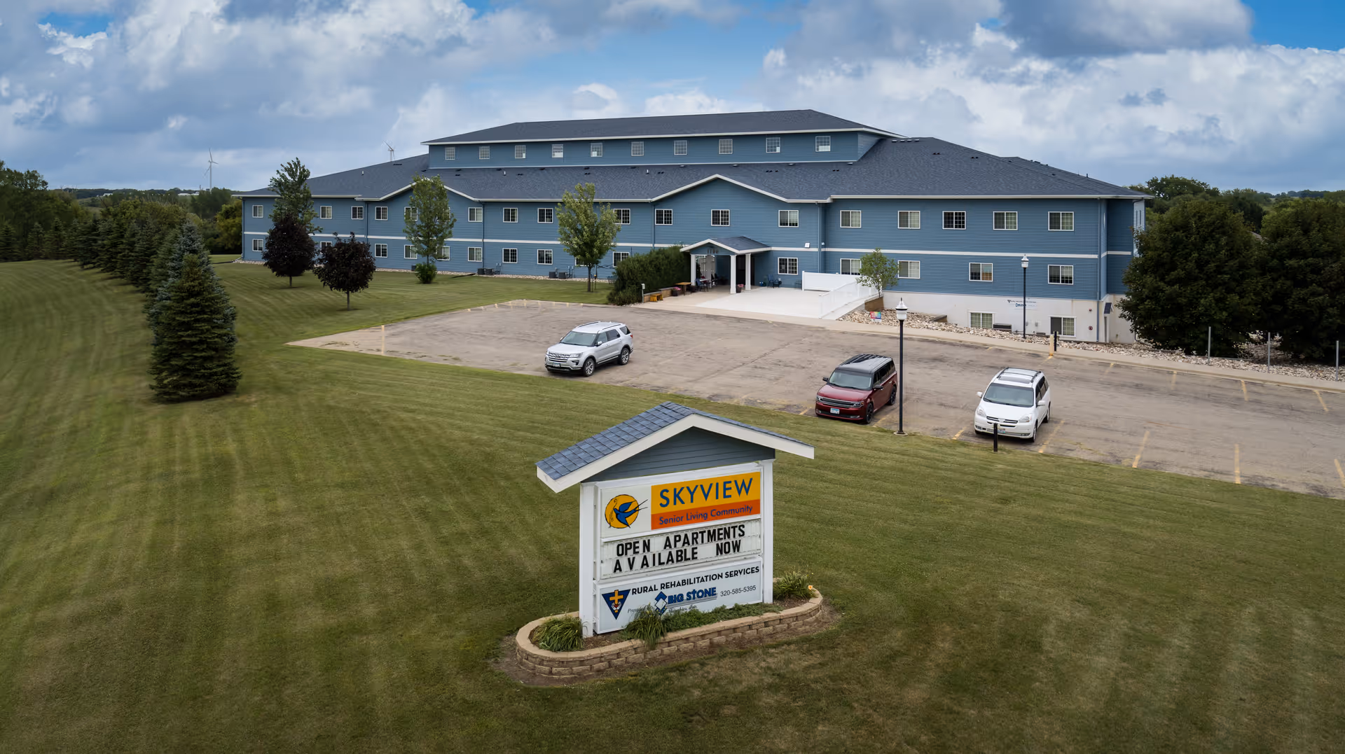 A large three-story blue senior living facility building with multiple windows and a main entrance with a small covered porch. In front of the building is a parking lot with three cars parked. A sign in the foreground reads 'Skyview Senior Living Community Open Apartments Available Now' with additional information about rehabilitation services. The building is surrounded by a well-maintained grassy area with several trees and a partly cloudy sky overhead.