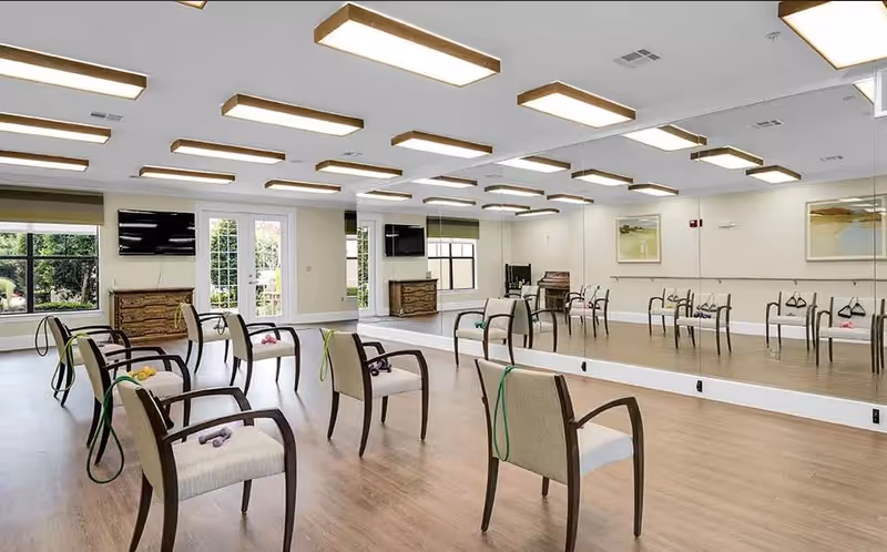 A spacious exercise or activity room with wooden flooring, several chairs arranged with resistance bands and small weights on them, large wall mirrors, two flat-screen TVs mounted on the walls, and windows letting in natural light.