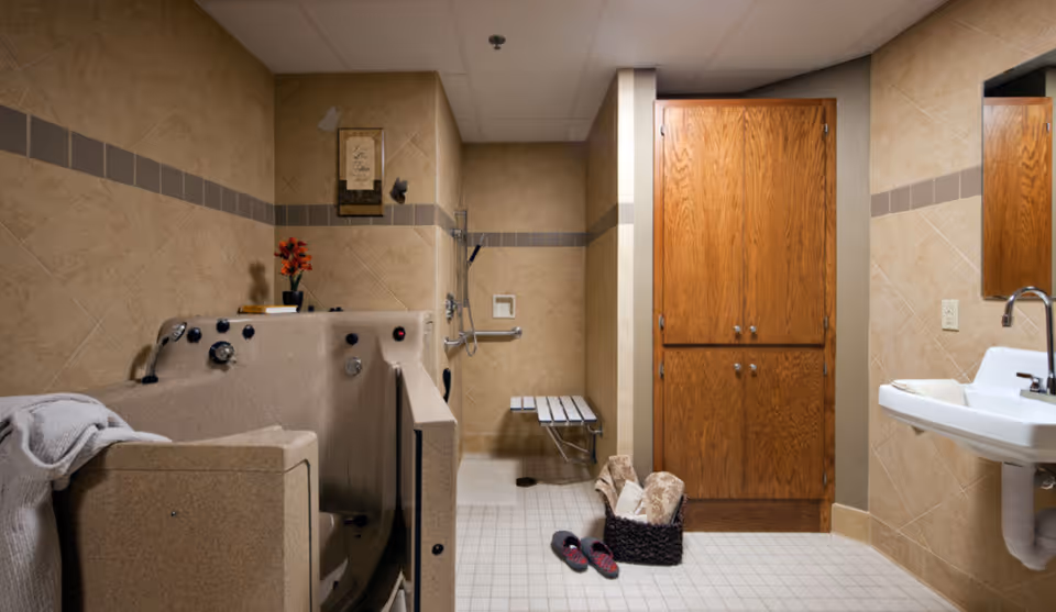 A bathroom with beige tiled walls and floor, featuring a walk-in bathtub with controls, a wall-mounted shower with a foldable bench, a wooden cabinet, a white sink with a towel, a basket with rolled towels, and a pair of slippers on the floor.
