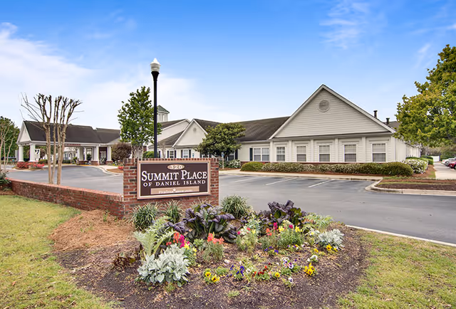 Exterior view of Summit Place of Daniel Island senior living facility with a brick sign in front surrounded by a flower bed and a parking area beside the building under a blue sky.